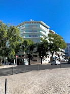 A multi-story residential building with pastel green and white colors surrounded by lush trees. A clear blue sky forms the backdrop, and a cobblestone sidewalk lines the street in front. Several parked cars are visible along the road, along with some street signs and a small advertisement board.