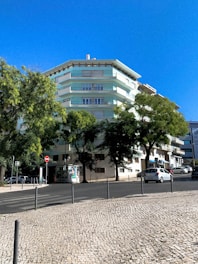 A multi-story residential building with pastel green and white colors surrounded by lush trees. A clear blue sky forms the backdrop, and a cobblestone sidewalk lines the street in front. Several parked cars are visible along the road, along with some street signs and a small advertisement board.