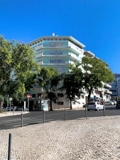 A multi-story residential building with pastel green and white colors surrounded by lush trees. A clear blue sky forms the backdrop, and a cobblestone sidewalk lines the street in front. Several parked cars are visible along the road, along with some street signs and a small advertisement board.