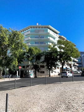 A multi-story residential building with pastel green and white colors surrounded by lush trees. A clear blue sky forms the backdrop, and a cobblestone sidewalk lines the street in front. Several parked cars are visible along the road, along with some street signs and a small advertisement board.