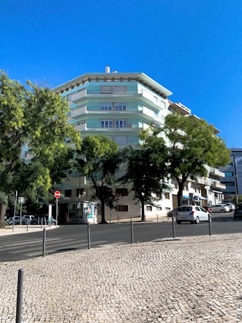 A multi-story residential building with pastel green and white colors surrounded by lush trees. A clear blue sky forms the backdrop, and a cobblestone sidewalk lines the street in front. Several parked cars are visible along the road, along with some street signs and a small advertisement board.