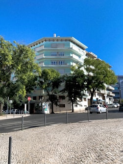 A multi-story residential building with pastel green and white colors surrounded by lush trees. A clear blue sky forms the backdrop, and a cobblestone sidewalk lines the street in front. Several parked cars are visible along the road, along with some street signs and a small advertisement board.