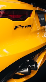 The image features the rear section of a bright yellow Jaguar F-Type car, showcasing its sleek tail light design and dual exhaust pipes. The car's logo is prominently displayed on the back, and the photo captures the glossy finish of the vehicle.