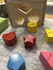 A wooden shape-sorting toy featuring cut-out spaces and various colored geometric wooden blocks labeled with their respective shapes. The blocks include a red heart, a red star, a blue pentagon, a yellow hexagon, a pink triangle, and a yellow section. These objects are placed on a textured surface next to the wooden sorter.