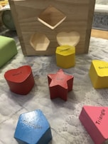 A wooden shape-sorting toy featuring cut-out spaces and various colored geometric wooden blocks labeled with their respective shapes. The blocks include a red heart, a red star, a blue pentagon, a yellow hexagon, a pink triangle, and a yellow section. These objects are placed on a textured surface next to the wooden sorter.