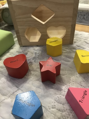 A wooden shape-sorting toy featuring cut-out spaces and various colored geometric wooden blocks labeled with their respective shapes. The blocks include a red heart, a red star, a blue pentagon, a yellow hexagon, a pink triangle, and a yellow section. These objects are placed on a textured surface next to the wooden sorter.