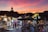 A bustling marketplace at sunset, with vibrant stalls selling colorful woven baskets and textiles. People are walking through the market, browsing the merchandise. The silhouette of a traditional tower is visible against a dramatic sky filled with hues of pink, orange, and purple.