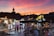 A bustling marketplace at sunset, with vibrant stalls selling colorful woven baskets and textiles. People are walking through the market, browsing the merchandise. The silhouette of a traditional tower is visible against a dramatic sky filled with hues of pink, orange, and purple.