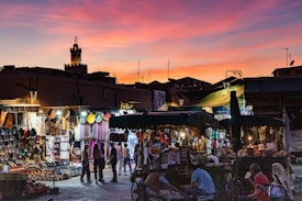 A bustling marketplace at sunset, with vibrant stalls selling colorful woven baskets and textiles. People are walking through the market, browsing the merchandise. The silhouette of a traditional tower is visible against a dramatic sky filled with hues of pink, orange, and purple.