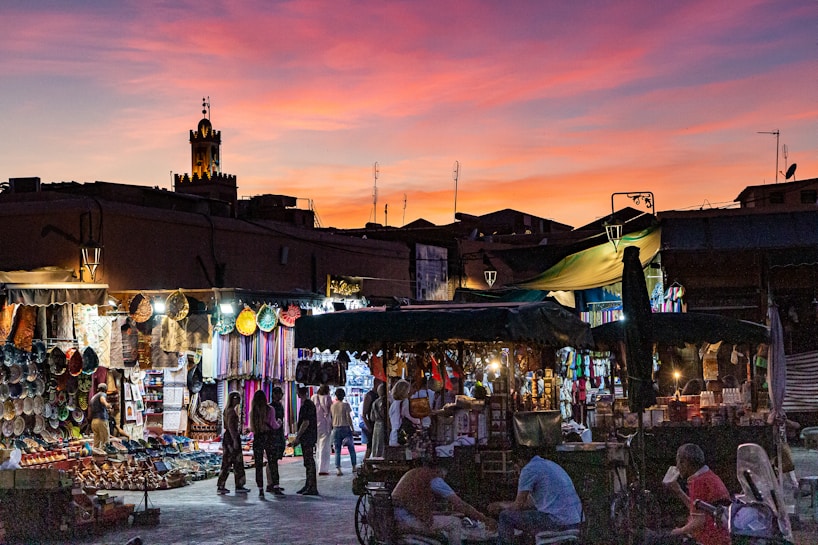 A bustling marketplace at sunset, with vibrant stalls selling colorful woven baskets and textiles. People are walking through the market, browsing the merchandise. The silhouette of a traditional tower is visible against a dramatic sky filled with hues of pink, orange, and purple.