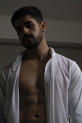 A man with short dark hair, facial hair, and a contemplative expression stands against a neutral background. He is wearing an open white shirt, revealing a well-defined torso with visible abs. The lighting casts subtle shadows across his face and body, creating a dramatic effect.