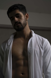A man with short dark hair, facial hair, and a contemplative expression stands against a neutral background. He is wearing an open white shirt, revealing a well-defined torso with visible abs. The lighting casts subtle shadows across his face and body, creating a dramatic effect.
