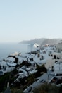 White buildings with blue domed roofs are clustered on a hillside with a view of the sea. People are gathered on a terrace, enjoying the scenic view. Rocky, mountainous terrain can be seen in the background.