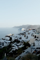 White buildings with blue domed roofs are clustered on a hillside with a view of the sea. People are gathered on a terrace, enjoying the scenic view. Rocky, mountainous terrain can be seen in the background.