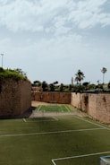 An outdoor sports complex featuring two soccer fields made of artificial turf, enclosed by high, historic stone walls with greenery growing on top. In the background, there are palm trees and additional greenery visible, under a sky with scattered clouds.