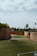 An outdoor sports complex featuring two soccer fields made of artificial turf, enclosed by high, historic stone walls with greenery growing on top. In the background, there are palm trees and additional greenery visible, under a sky with scattered clouds.