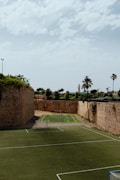 An outdoor sports complex featuring two soccer fields made of artificial turf, enclosed by high, historic stone walls with greenery growing on top. In the background, there are palm trees and additional greenery visible, under a sky with scattered clouds.