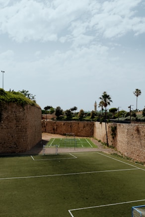 An outdoor sports complex featuring two soccer fields made of artificial turf, enclosed by high, historic stone walls with greenery growing on top. In the background, there are palm trees and additional greenery visible, under a sky with scattered clouds.
