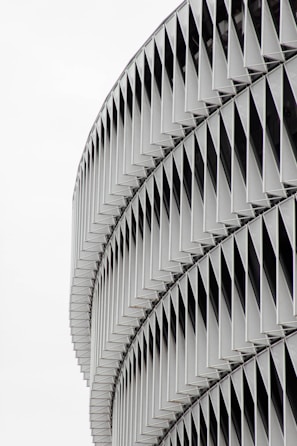 A modern architectural facade with a series of repetitive geometric patterns, consisting of overlapping metal panels that create a visually striking and intricate design. The panels are arranged in a vertical alignment with slight curvature.