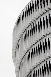 A modern architectural facade with a series of repetitive geometric patterns, consisting of overlapping metal panels that create a visually striking and intricate design. The panels are arranged in a vertical alignment with slight curvature.