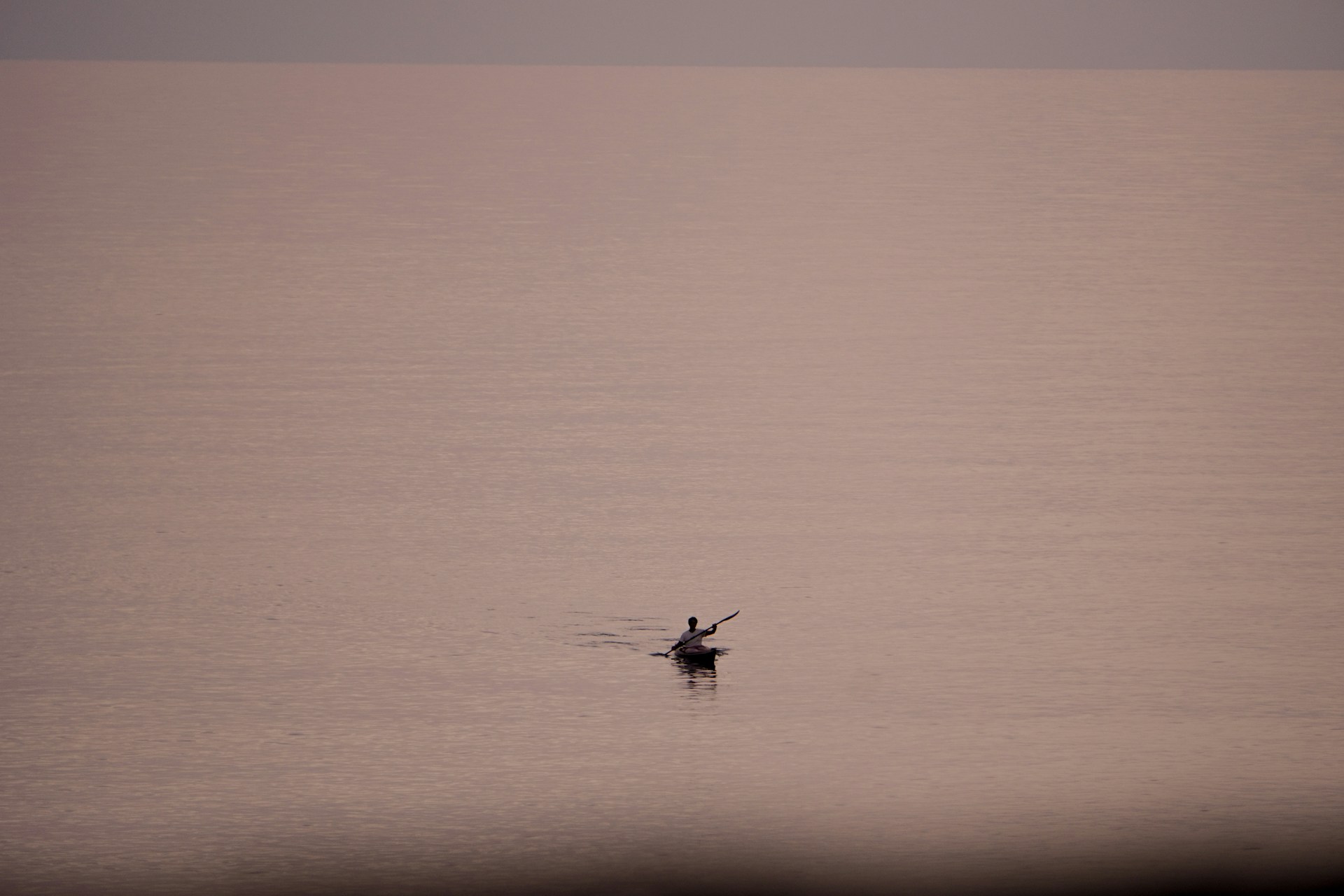 A lone kayaker paddles across a vast, calm body of water under a serene sky. The water and sky possess a soft, muted color palette, creating a tranquil scene.