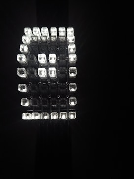A geometric arrangement of multiple small, rectangular light cubes emitting a bright, white light against a dark background. The cubes are organized in a grid-like, three-dimensional structure, creating a striking visual effect.
