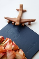 A wooden airplane model is placed on top of a blue document or diploma cover. In the foreground, there is a bouquet of peach and pink roses. The document has an embossed seal with Russian text.