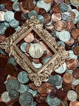 A collection of various coins is scattered across the surface, with a vintage, ornate golden frame placed centrally around one of the coins. The coins vary in color and denomination, with some depicting designs and figures.