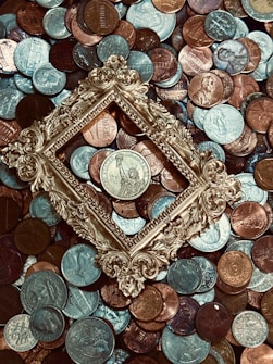 A collection of various coins is scattered across the surface, with a vintage, ornate golden frame placed centrally around one of the coins. The coins vary in color and denomination, with some depicting designs and figures.