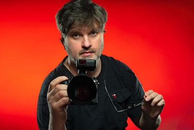 A person holding a camera with a large lens and a flash attachment, standing against a vivid red background. The individual has medium-length hair and is wearing a black shirt.