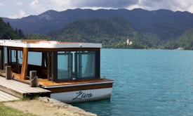 A boat named 'Živa' is docked at a wooden pier against a backdrop of turquoise water and lush green mountains. In the distance, a small white church with a tower is nestled on an island. The sky is partly cloudy, adding a serene atmosphere to the scenic view.