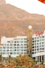 A modern hotel building with a distinct cylindrical tower, adorned with a spherical white structure on top. The facade features multiple windows with rounded arches, and a large sign with the word 'Enjoy' in red letters is prominently displayed. In the foreground, lush green palm trees and leafy bushes add a touch of nature. The backdrop consists of imposing rocky cliffs under a clear sky.