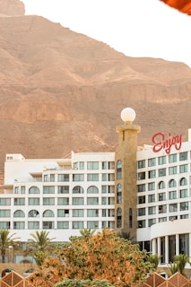 A modern hotel building with a distinct cylindrical tower, adorned with a spherical white structure on top. The facade features multiple windows with rounded arches, and a large sign with the word 'Enjoy' in red letters is prominently displayed. In the foreground, lush green palm trees and leafy bushes add a touch of nature. The backdrop consists of imposing rocky cliffs under a clear sky.