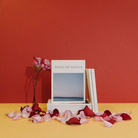 A stack of books titled 'Song of Songs' is accompanied by two pink roses in a vase. Rose petals are scattered on a yellow surface against a red backdrop.