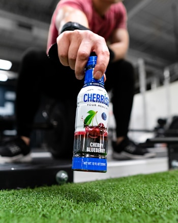 A person holding a bottle labeled 'CHERRiSH Cherry Blueberry' in a gym setting, with their hand extended toward the camera. The focus is on the bottle, while gym equipment and artificial grass can be seen in the background.