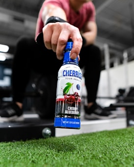 A person holding a bottle labeled 'CHERRiSH Cherry Blueberry' in a gym setting, with their hand extended toward the camera. The focus is on the bottle, while gym equipment and artificial grass can be seen in the background.