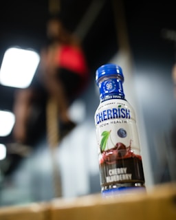 A bottle of Cheribundi cherry blueberry juice is prominently placed in the foreground, with clear branding and design elements visible. In the blurred background, a person appears to be climbing a rope, suggesting an active environment.
