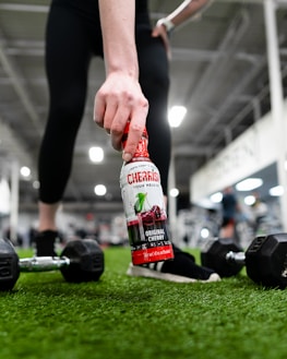 A hand is holding a bottle of cherry-flavored beverage on a turf surface in a gym setting. Dumbbells are placed on the ground nearby, and the blurred background indicates other gym equipment and people.