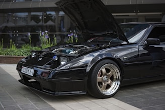 A sleek black sports car is parked with its hood open to reveal the engine. The car is situated on a tiled surface with reflective glass windows and plants in the background, showcasing purple flowers. The front wheel features a stylish chrome rim and GT branding.