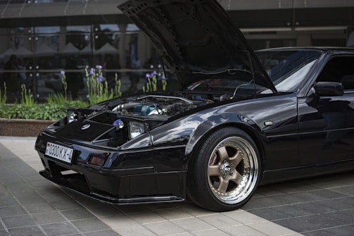 A sleek black sports car is parked with its hood open to reveal the engine. The car is situated on a tiled surface with reflective glass windows and plants in the background, showcasing purple flowers. The front wheel features a stylish chrome rim and GT branding.