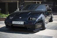 A sleek, black sports car is parked on a tiled surface, reflecting light off its polished exterior. The vehicle features distinct, angular headlights and a low profile with wide tires. A license plate with Cyrillic characters is visible, indicating its registration.