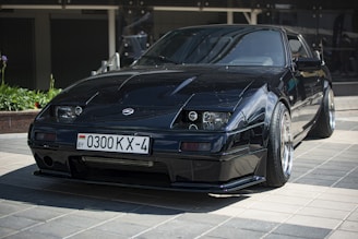 A sleek, black sports car is parked on a tiled surface, reflecting light off its polished exterior. The vehicle features distinct, angular headlights and a low profile with wide tires. A license plate with Cyrillic characters is visible, indicating its registration.