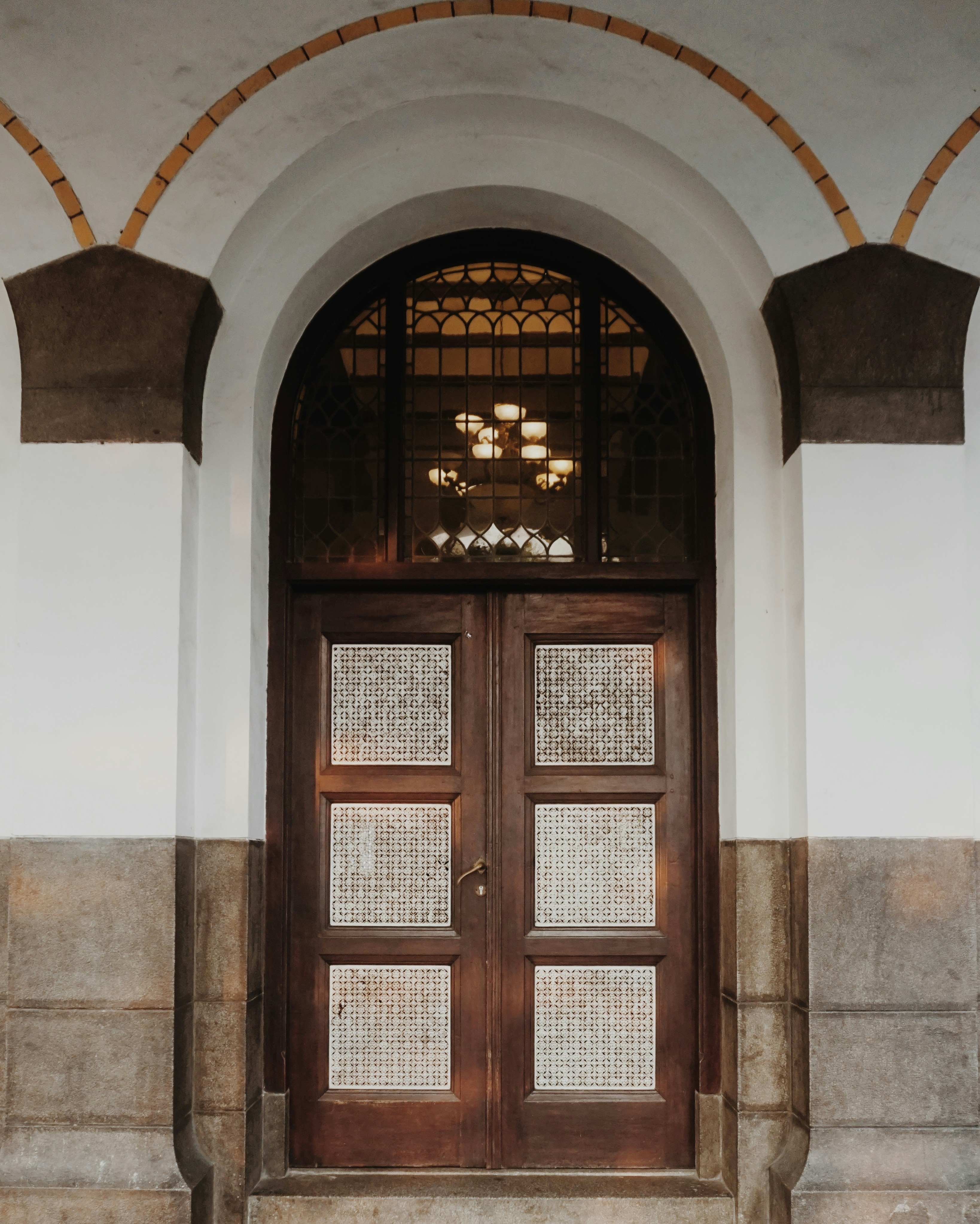 An ornate wooden door with glass panels featuring a decorative grid pattern stands beneath a large arched entryway. The arch is framed by light gray and dark brown stonework, adding a feeling of elegance. The door is set in a white wall and there are intricate glass panes above the door.
