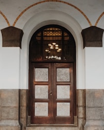 An ornate wooden door with glass panels featuring a decorative grid pattern stands beneath a large arched entryway. The arch is framed by light gray and dark brown stonework, adding a feeling of elegance. The door is set in a white wall and there are intricate glass panes above the door.