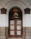 An ornate wooden door with glass panels featuring a decorative grid pattern stands beneath a large arched entryway. The arch is framed by light gray and dark brown stonework, adding a feeling of elegance. The door is set in a white wall and there are intricate glass panes above the door.