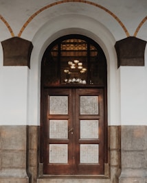 An ornate wooden door with glass panels featuring a decorative grid pattern stands beneath a large arched entryway. The arch is framed by light gray and dark brown stonework, adding a feeling of elegance. The door is set in a white wall and there are intricate glass panes above the door.