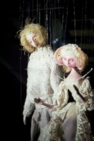 Two marionette puppets with elaborate costumes and intricate detailing. One puppet has blond curly hair and is wearing a textured knitted sweater, while the other has pinkish hair adorned with flowers and is dressed in an elegant lacy dress. Both are suspended by strings against a dark background, giving them a lifelike appearance.