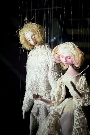 Two marionette puppets with elaborate costumes and intricate detailing. One puppet has blond curly hair and is wearing a textured knitted sweater, while the other has pinkish hair adorned with flowers and is dressed in an elegant lacy dress. Both are suspended by strings against a dark background, giving them a lifelike appearance.
