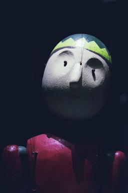A close-up of a wooden marionette puppet with a round head, small facial features, and a painted green and yellow zigzag pattern resembling a cap. The puppet is dressed in a red outfit, and strings are visible, suggesting it is a string puppet. The lighting is dim, casting shadows and giving a dramatic effect.