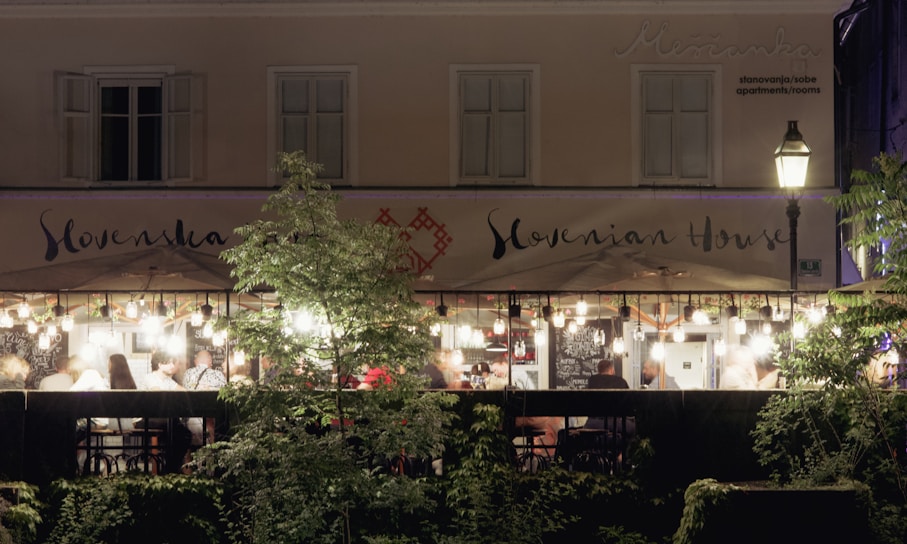 An outdoor dining area at a restaurant is brightly illuminated with strings of bulbs, creating a warm and inviting atmosphere. Patrons are seen sitting under umbrellas, enjoying their meals. The facade of a building in the background displays the sign 'Slovenian House'. A tree and shrubs partially obscure the view, adding a touch of greenery.